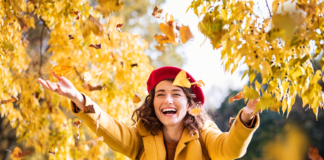 Auf dem Bild ist eine lebendige Frau im Herbst zu sehen, die in einem Park steht. Sie trägt warme Herbstkleidung und wirft mit einem strahlenden Lächeln bunte Herbstblätter in die Luft. Der goldene Sonnenschein betont ihre lebhafte Energie, während die fallenden Blätter die Magie und Freude des Herbstes einfangen.